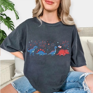 Puede incluir: Camiseta gris oscuro con una fila de dinosaurios de dibujos animados en azul y rojo, con fuegos artificiales y una bandera estadounidense. Los dinosaurios incluyen un T-Rex azul, un Estegosaurio azul, un Brontosaurio azul y un Triceratops rojo.