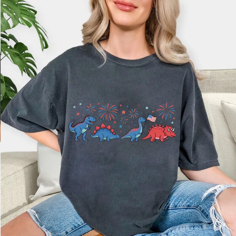 Puede incluir: Camiseta gris oscuro con una fila de dinosaurios de dibujos animados en azul y rojo, con fuegos artificiales y una bandera estadounidense. Los dinosaurios incluyen un T-Rex azul, un Estegosaurio azul, un Brontosaurio azul y un Triceratops rojo.