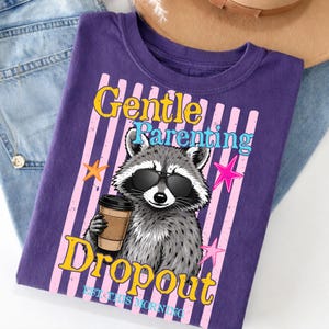 Op de afbeelding: Paarse t-shirt met een wasbeer met een zonnebril en een koffiekopje. Het shirt heeft de woorden "Gentle Parenting Dropout" in geel en blauw, met roze en witte strepen.