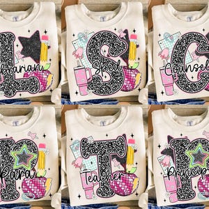 May include: Cream-colored sweatshirts with various school-themed designs. Each sweatshirt features a large letter, the word for a school position, and colorful graphics including a pencil, apple, and disco ball. The sweatshirts are perfect for teachers and school staff.