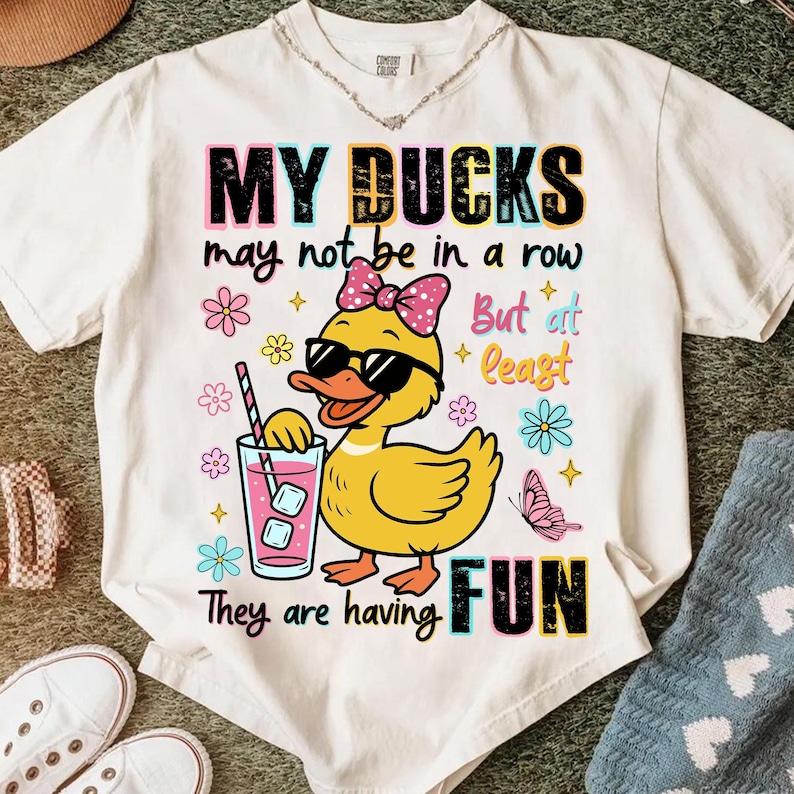May include: White t-shirt with a graphic of a yellow duck wearing sunglasses and a pink bow, holding a drink. Text reads "MY DUCKS may not be in a row But at least They are having FUN."