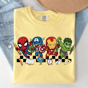 May include: Yellow t-shirt featuring cartoon illustrations of Spider-Man, Captain America, Iron Man, and the Hulk, each holding a pencil. The design includes school-themed elements like a globe, books, and a clock.