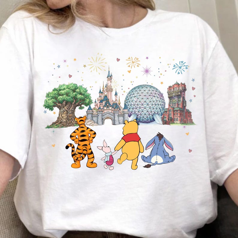 May include: White t-shirt featuring a colorful graphic of Winnie the Pooh and friends in front of iconic theme park landmarks. The design includes a tiger, piglet, bear, and donkey, with fireworks and a tree.