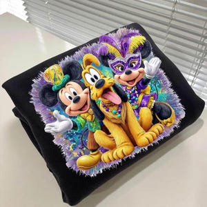 K&ouml;nnte beinhalten: Schwarzes Sweatshirt mit Mickey Mouse, Minnie Mouse und Pluto in festlicher Mardi-Gras-Kleidung. Mickey tr&auml;gt einen gr&uuml;nen und goldenen Hut und Pluto eine Perlenkette. Minnie tr&auml;gt eine lila Maske und einen Federkopfschmuck.
