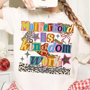 Pu&ograve; includere: T-shirt color crema con la scritta multicolore "Motherhood is Kingdom Work". Il design include stampa leopardata, stelle e un fiocco. Il testo "She is strong...Proverbi 31:25-27" &egrave; stampato in basso.