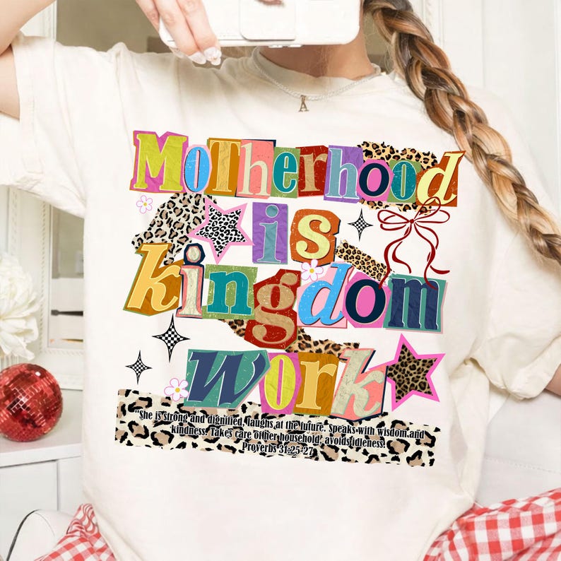 Pu&ograve; includere: T-shirt color crema con la scritta multicolore "Motherhood is Kingdom Work". Il design include stampa leopardata, stelle e un fiocco. Il testo "She is strong...Proverbi 31:25-27" &egrave; stampato in basso.