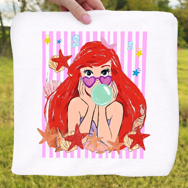 May include: White square towel with a cartoon mermaid illustration. The mermaid has red hair, heart-shaped sunglasses, and a bubble. The design includes pink and white stripes, starfish, seashells, and bubbles.