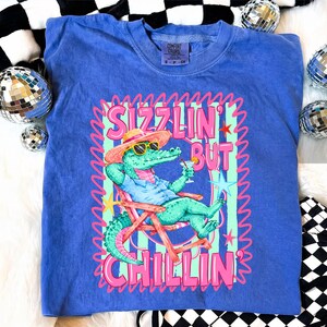 May include: A blue Comfort Colors t-shirt featuring a cartoon alligator in sunglasses, a hat, and a drink, relaxing in a beach chair. The shirt has the text "Sizzlin' But Chillin'" in pink and green.