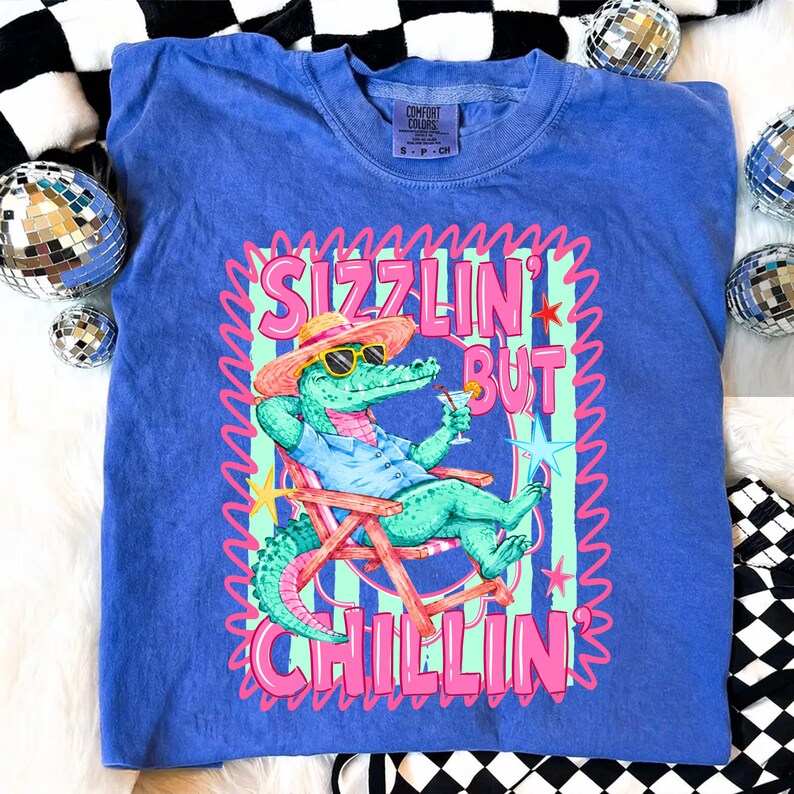 May include: A blue Comfort Colors t-shirt featuring a cartoon alligator in sunglasses, a hat, and a drink, relaxing in a beach chair. The shirt has the text "Sizzlin' But Chillin'" in pink and green.
