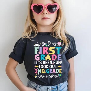 Puede incluir: Una camiseta gris oscuro con el texto "So long First Grade, It's Been Fun! Look Out 2nd Grade, Here I Come!" en letras coloridas. La camiseta presenta un gorro de graduaci&oacute;n y otros elementos decorativos.