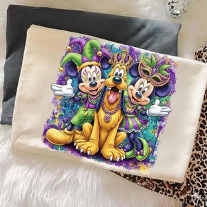 May include: Off-white t-shirt featuring a colorful graphic of Mickey Mouse, Minnie Mouse, and Pluto in Mardi Gras attire. Mickey and Minnie wear jester hats and masks, while Pluto has a crown. The design includes vibrant purples, greens, and blues.