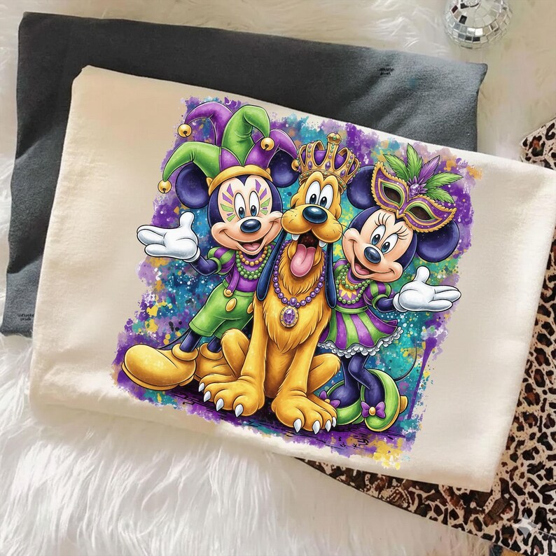 May include: Off-white t-shirt featuring a colorful graphic of Mickey Mouse, Minnie Mouse, and Pluto in Mardi Gras attire. Mickey and Minnie wear jester hats and masks, while Pluto has a crown. The design includes vibrant purples, greens, and blues.