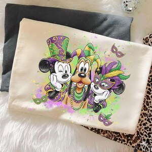 May include: A cream-colored pillowcase featuring a colorful illustration of Mickey Mouse, Goofy, and another Mickey Mouse character in Mardi Gras attire. The characters are wearing purple, green, and gold outfits and masks. The design includes confetti and decorative masks.