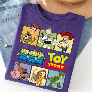 Puede incluir: Una camiseta morada con personajes de la pel&iacute;cula animada "Toy Story". El dise&ntilde;o incluye im&aacute;genes de Woody, Buzz Lightyear, Jessie, los Aliens y otros personajes. Las palabras "Toy Story" son en amarillo y rojo.