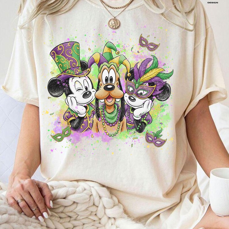May include: Cream-colored t-shirt featuring a colorful graphic of Mickey Mouse, Goofy, and another Mickey Mouse character in Mardi Gras attire. The characters are wearing masks, hats, and beads. The design includes purple, green, and gold accents.