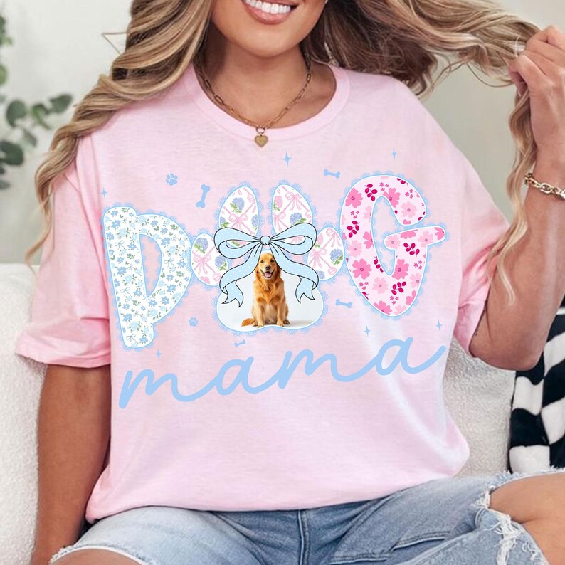 May include: A light pink t-shirt with the words "Dog Mama" in light blue script. The word "Dog" is designed with a paw print, a bow, and a golden retriever image. The letter "G" is decorated with pink flowers.