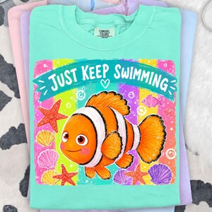 K&ouml;nnte beinhalten: Ein t&uuml;rkisfarbenes Comfort Colors T-Shirt mit einer bunten Grafik eines Cartoon-Clownfischs und dem Text "Just Keep Swimming". Das Design umfasst Seesterne und Muscheln. Das T-Shirt ist gefaltet.