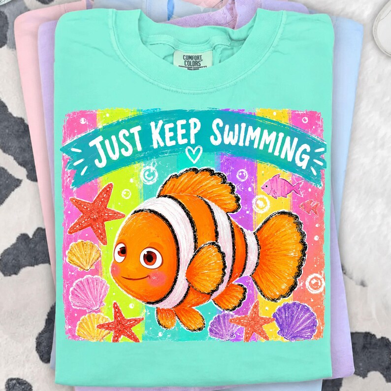 K&ouml;nnte beinhalten: Ein t&uuml;rkisfarbenes Comfort Colors T-Shirt mit einer bunten Grafik eines Cartoon-Clownfischs und dem Text "Just Keep Swimming". Das Design umfasst Seesterne und Muscheln. Das T-Shirt ist gefaltet.