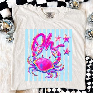 May include: White t-shirt with a colorful crab graphic and the word "Oh". The crab is pink, blue, and yellow, set against a light blue striped background. Three pink stars are also featured.