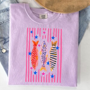 Pode incluir: Uma camiseta lil&aacute;s Comfort Colors com um design de peixes coloridos. O gr&aacute;fico inclui tr&ecirc;s peixes com padr&otilde;es diferentes, listras verticais rosa e estrelas azuis. A camiseta tem gola redonda e mangas curtas.