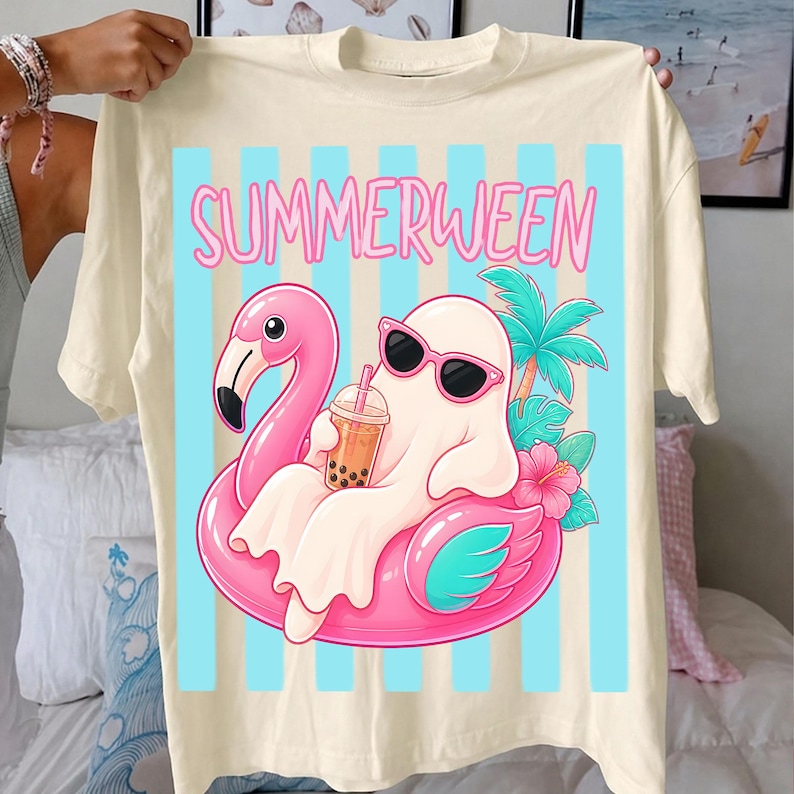 K&ouml;nnte beinhalten: Cremefarbenes T-Shirt mit dem Wort "SUMMERWEEN" in Pink. Die Grafik zeigt einen Geist mit Sonnenbrille, der auf einem rosa Flamingo-Schwimmer liegt und ein Getr&auml;nk h&auml;lt. Blaue vertikale Streifen im Hintergrund.