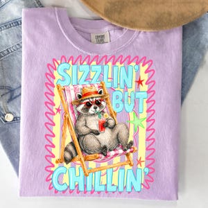 May include: A light purple t-shirt featuring a raccoon in sunglasses and a straw hat, relaxing in a beach chair with a drink. The shirt has the words "Sizzlin' But Chillin'" in a colorful design.
