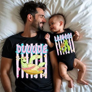 May include: Matching black t-shirts and onesie with a cartoon turtle design. The adult shirt says "DUUUUDE" in pink and blue, while the baby's onesie says "LIL DUDE". Both feature a green turtle on a yellow base with pink and white striped backgrounds.