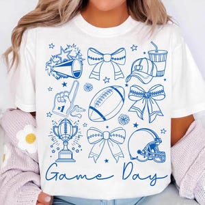 May include: White t-shirt with a blue graphic design featuring football-themed elements. The design includes a football, helmet, bows, a trophy, and the words "Game Day". Stars and other shapes are scattered around the design.
