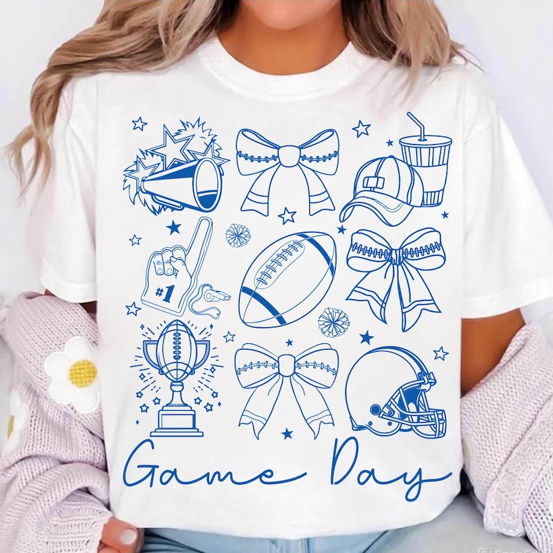 May include: White t-shirt with a blue graphic design featuring football-themed elements. The design includes a football, helmet, bows, a trophy, and the words "Game Day". Stars and other shapes are scattered around the design.