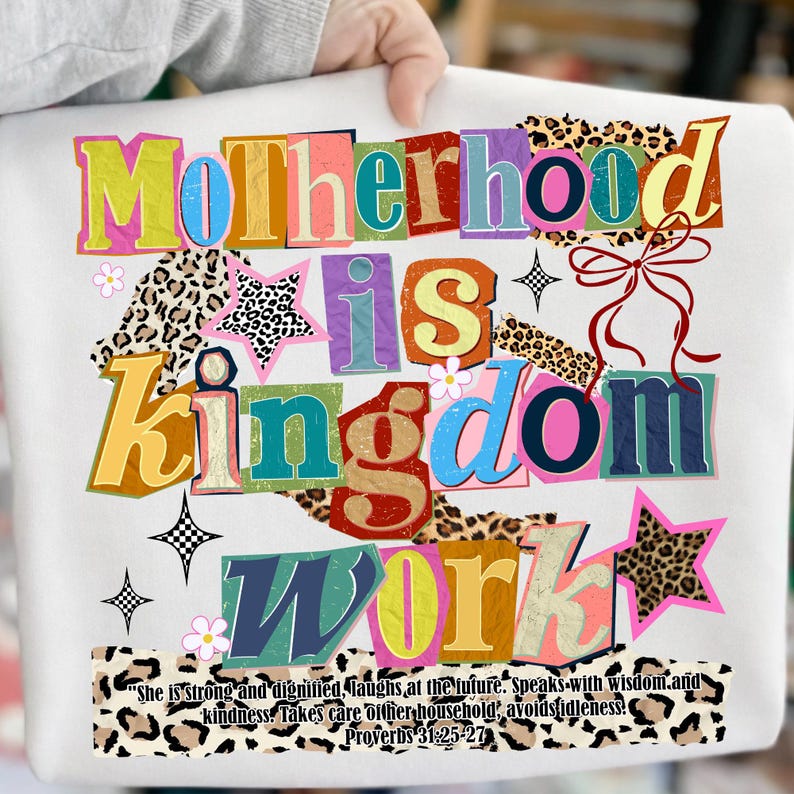 Pu&ograve; includere: Felpa bianca con la scritta "Motherhood is Kingdom Work" in lettere colorate sovrapposte. Il design include dettagli floreali e animalier, con una citazione di Proverbi 31:25-27 in basso.