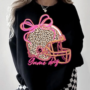 Könnte beinhalten: Schwarzes Sweatshirt mit einem Footballhelm-Design. Der Helm hat ein Leopardenmuster, eine rosa Schleife und goldene Umrisse. Die Worte "Game day" sind in rosa Schrift darunter geschrieben.