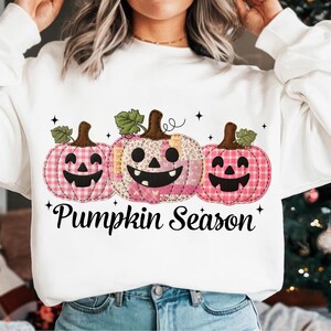 May include: White sweatshirt featuring three smiling pink pumpkins with green leaves and brown stems. The pumpkins have different patterns, including gingham and floral designs. The text "Pumpkin Season" is printed below the pumpkins.