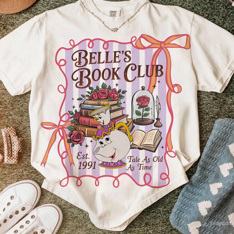 May include: White t-shirt with a graphic for "Belle's Book Club." The design includes books, roses, a glass-encased rose, and the text "Tale As Old As Time." The shirt also has the text "Est. 1991."