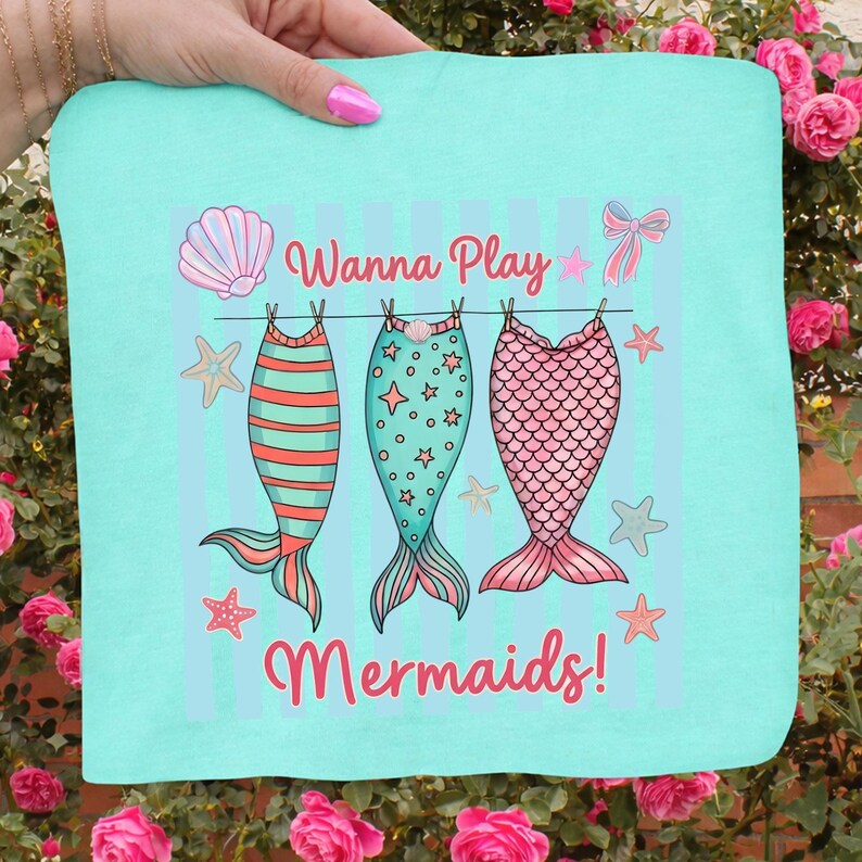 May include: A turquoise fabric with a whimsical design featuring three mermaid tails hanging on a clothesline. The tails are in shades of orange, teal, and pink, with the words "Wanna Play Mermaids!" in a playful font.