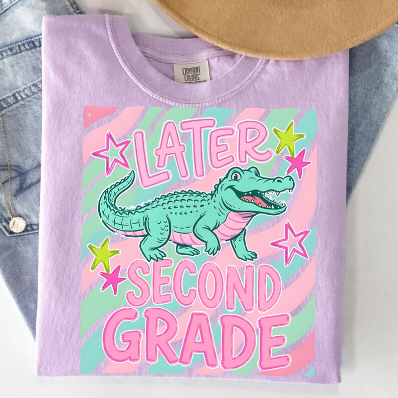 May include: A light purple t-shirt with a graphic of a teal alligator and the words "LATER SECOND GRADE" in pink. The design includes pink and green stars and a swirled background.