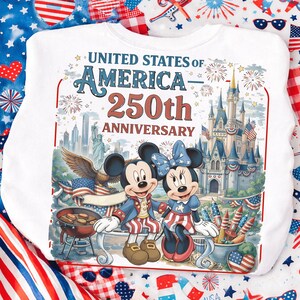 May include: White sweatshirt featuring Mickey and Minnie Mouse in patriotic attire, celebrating the 250th anniversary of the United States of America. The design includes fireworks, a castle, and the text "UNITED STATES OF AMERICA 250th ANNIVERSARY."