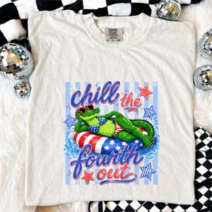 May include: Cream-colored t-shirt featuring a graphic of a green alligator wearing sunglasses and a bikini, lounging on a red, white, and blue inner tube. The text "Chill the Fourth Out" is displayed in blue and red script.