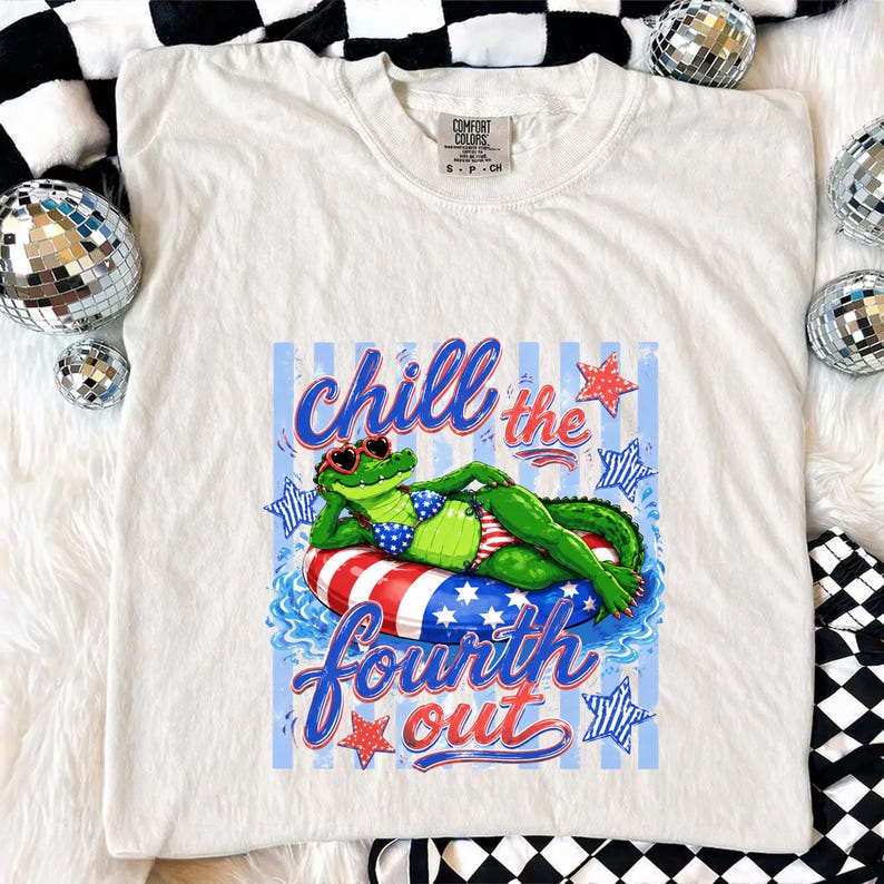 May include: Cream-colored t-shirt featuring a graphic of a green alligator wearing sunglasses and a bikini, lounging on a red, white, and blue inner tube. The text "Chill the Fourth Out" is displayed in blue and red script.