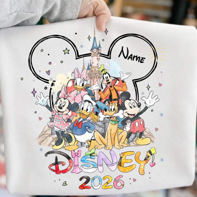 May include: White sweatshirt featuring a colorful Disney design with Mickey Mouse, Minnie Mouse, Donald Duck, Daisy Duck, Goofy, and Pluto. The word "Disney" is written in colorful letters, with "2026" below. A space is provided for a name.