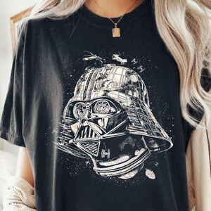 May include: Black t-shirt featuring a detailed graphic of Darth Vader's helmet, incorporating elements of space and starships. The design is in shades of white and gray, creating a striking contrast against the dark fabric. A unique and stylish graphic tee.