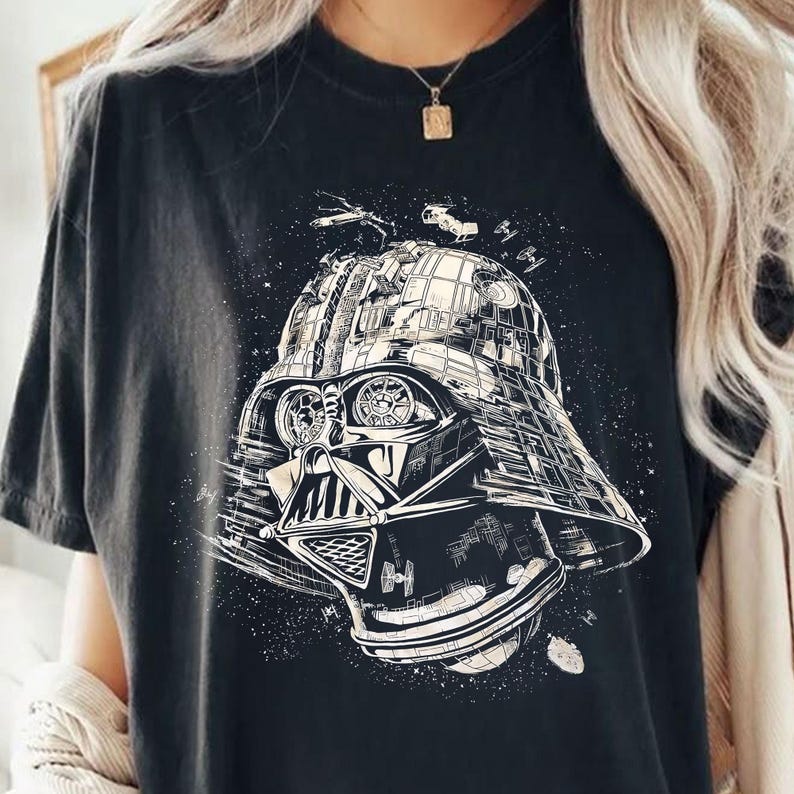 May include: Black t-shirt featuring a detailed graphic of Darth Vader's helmet, incorporating elements of space and starships. The design is in shades of white and gray, creating a striking contrast against the dark fabric. A unique and stylish graphic tee.