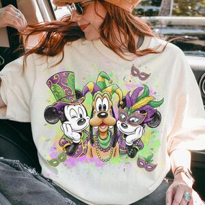 May include: Cream-colored sweatshirt featuring Mickey Mouse, Goofy, and another Mickey Mouse character in Mardi Gras attire. The characters wear purple, green, and gold outfits and masks. The design includes decorative masks and colorful paint splatters.