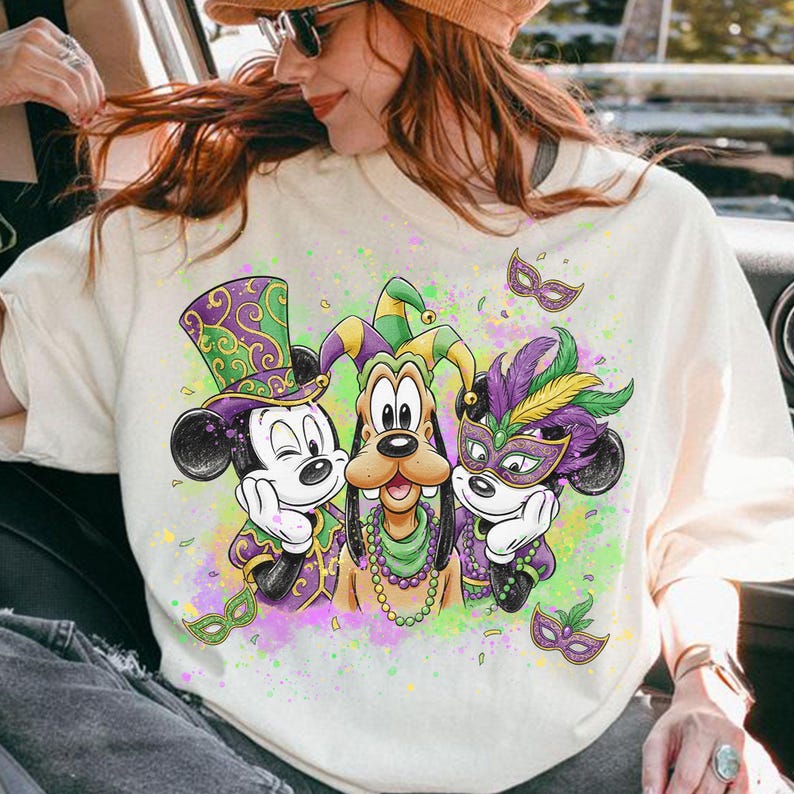 May include: Cream-colored sweatshirt featuring Mickey Mouse, Goofy, and another Mickey Mouse character in Mardi Gras attire. The characters wear purple, green, and gold outfits and masks. The design includes decorative masks and colorful paint splatters.