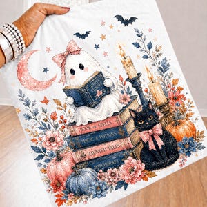 Peut inclure: Un article en tissu blanc illustré d'un fantôme avec un nœud rose lisant un livre intitulé Ghost Stories, assis sur une pile de livres nommés Book of Spells, Magic & Potions, Haunted Tales et Witchcraft, avec un chat noir, des citrouilles et des bougies.
