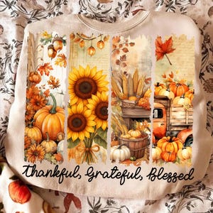 May include: Cream-colored sweatshirt with a fall-themed graphic. The design features pumpkins, sunflowers, and autumn leaves in shades of orange, yellow, and brown. The words "Thankful, Grateful, Blessed" are written in a script font.
