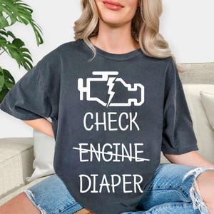 May include: A dark gray t-shirt with a white check engine light icon and the text "CHECK ENGINE DIAPER" with the word "ENGINE" crossed out.