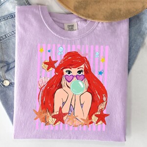 May include: A lilac t-shirt with a cartoon mermaid illustration. The mermaid has red hair, heart-shaped sunglasses, and a bubble. The design includes seashells, starfish, and bubbles against a pink striped background. The shirt has the text "Comfort Colors".