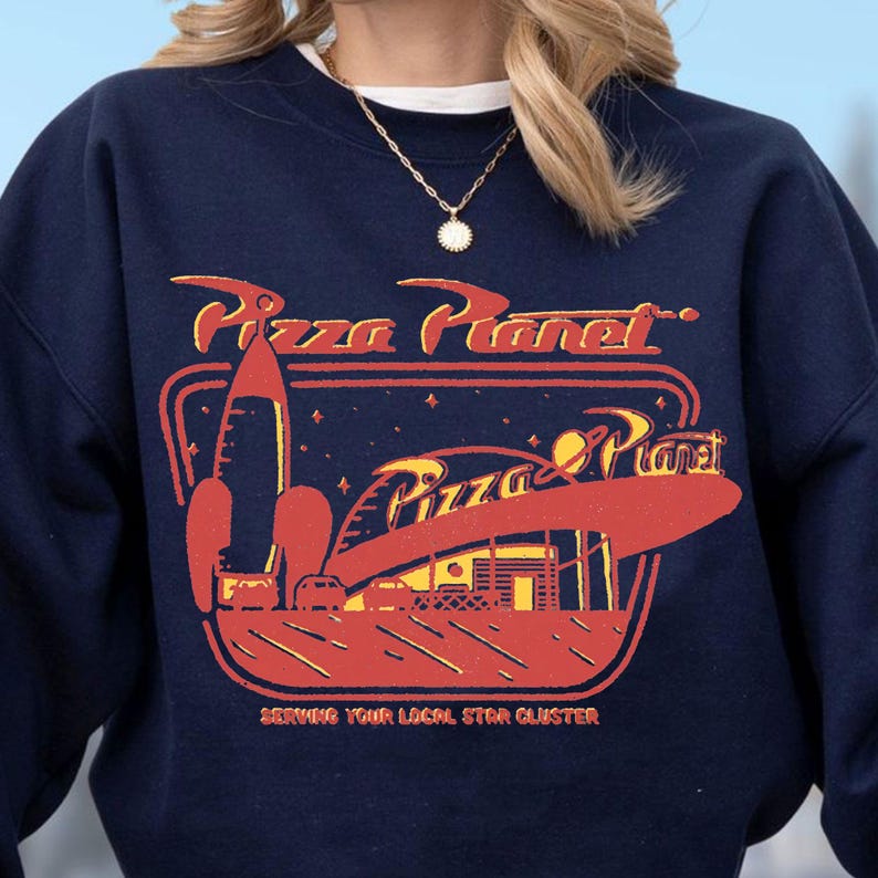 Puede incluir: Sudadera azul marino con un gr&aacute;fico retro de Pizza Planet en rojo y amarillo. El dise&ntilde;o incluye un cohete, un fondo tem&aacute;tico espacial y el texto "Pizza Planet" y "Serving Your Local Star Cluster."