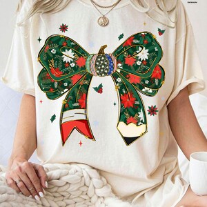 May include: Cream-colored t-shirt featuring a large, festive bow design. The bow is green with red floral and white accents, and a pumpkin-shaped center. The design includes gold detailing and is decorated with stars and holly.