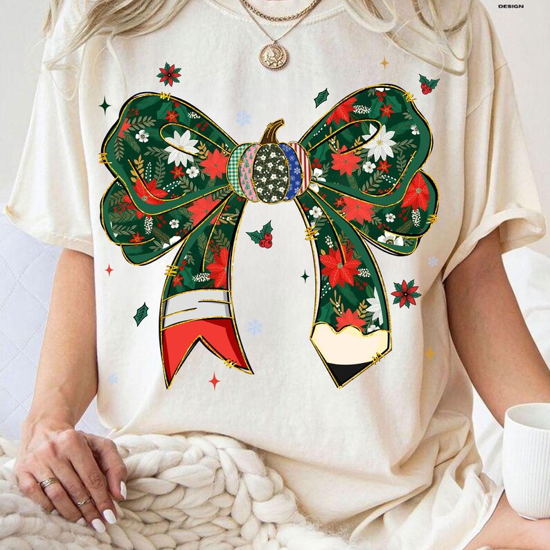 May include: Cream-colored t-shirt featuring a large, festive bow design. The bow is green with red floral and white accents, and a pumpkin-shaped center. The design includes gold detailing and is decorated with stars and holly.